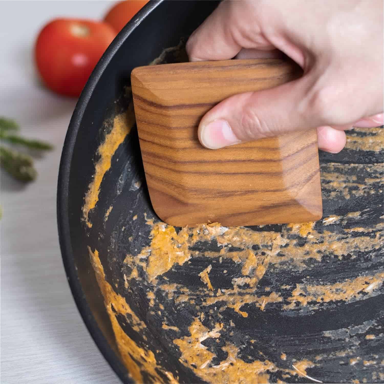 Alt: Hand using wooden scraper to clean black and gold culinary pot with food residue.