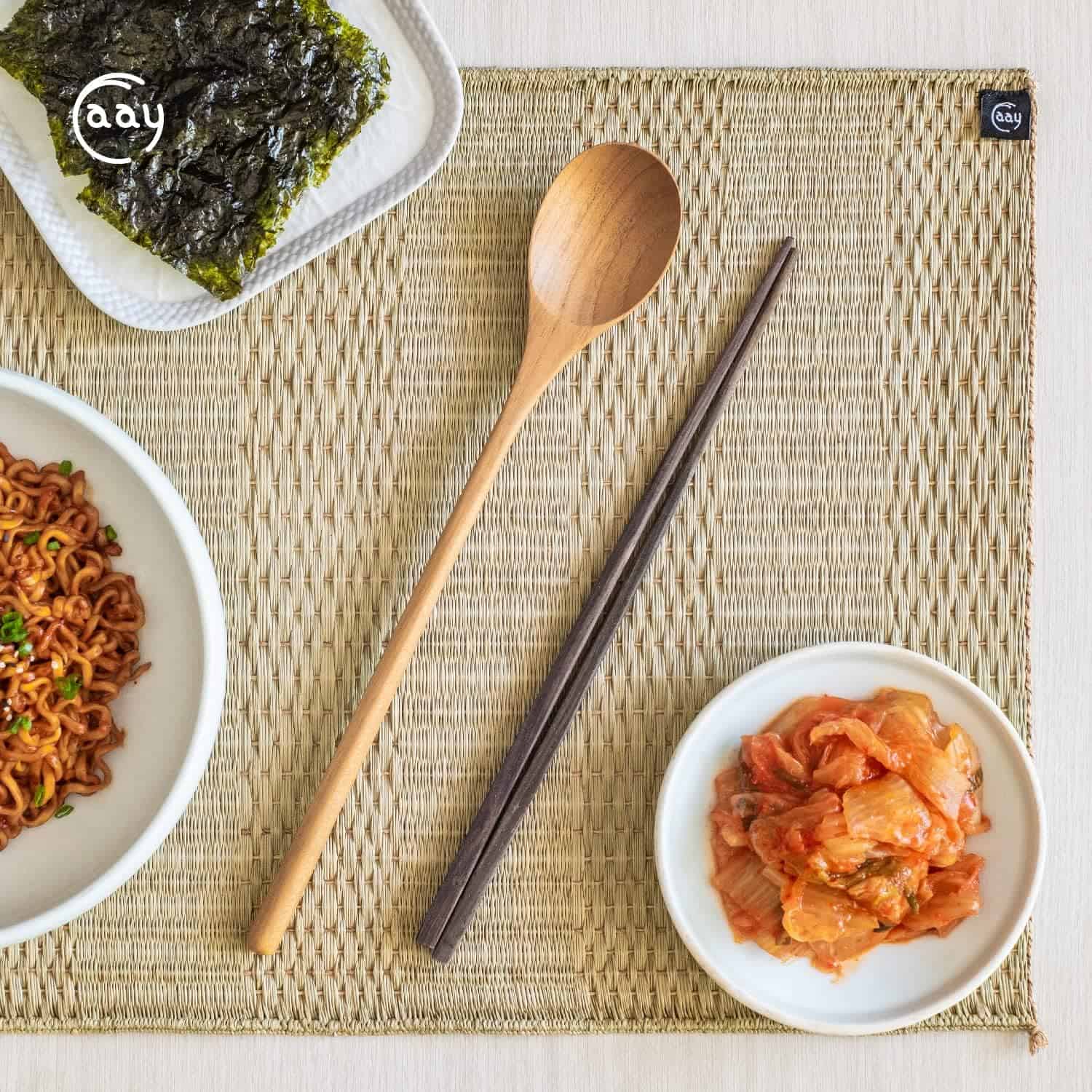 Delicious Korean pickled vegetables, seaweed, and noodles served with chopsticks and a spoon on woven placemat.
